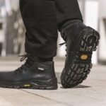 Une personne chaussée de bottes de travail Jallatte noires, dotées de semelles Vibram épaisses et robustes, marche sur une surface pavée. Les semelles des bottes arborent le logo Vibram en jaune. L'image met l'accent sur les chaussures de sécurité et le bas des jambes, à l'extérieur, à la lumière du jour.