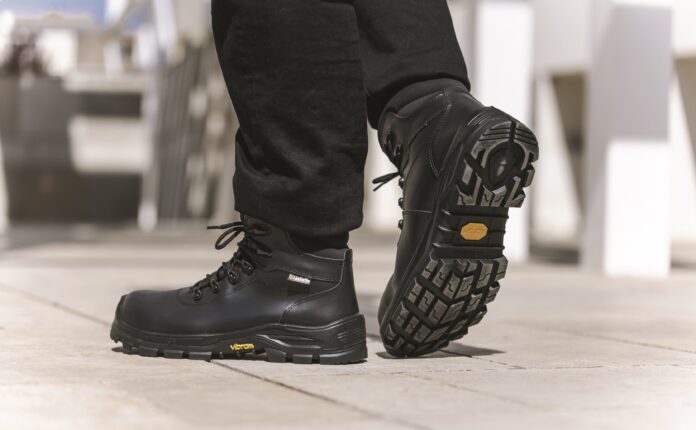 Une personne chaussée de bottes de travail Jallatte noires, dotées de semelles Vibram épaisses et robustes, marche sur une surface pavée. Les semelles des bottes arborent le logo Vibram en jaune. L'image met l'accent sur les chaussures de sécurité et le bas des jambes, à l'extérieur, à la lumière du jour.