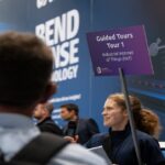 Une femme tient une pancarte violette sur laquelle on peut lire « Visites guidées – Visite n° 1 : Internet des objets industriel (IIoT) » tout en s’adressant aux participants d’un événement technologique. À l’arrière-plan, on aperçoit un stand consacré à la technologie et plusieurs personnes.