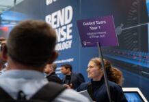 EuroBLECH 2026 ouvre la vente des billets visiteurs Une femme tient une pancarte violette sur laquelle on peut lire « Visites guidées – Visite n° 1 : Internet des objets industriel (IIoT) » tout en s’adressant aux participants d’un événement technologique. À l’arrière-plan, on aperçoit un stand consacré à la technologie et plusieurs personnes.