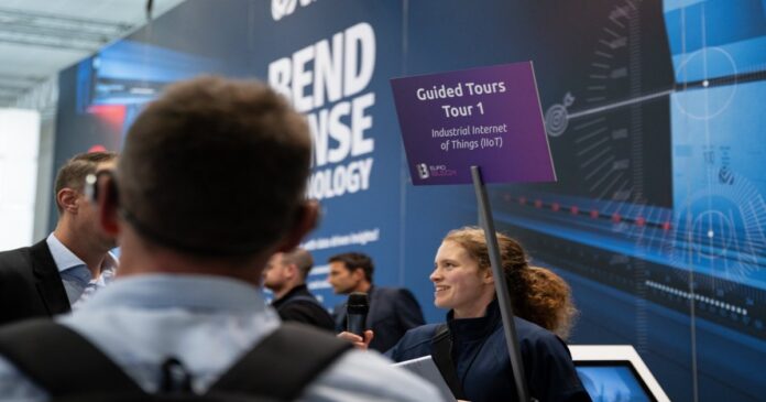 Une femme tient une pancarte violette sur laquelle on peut lire « Visites guidées – Visite n° 1 : Internet des objets industriel (IIoT) » tout en s’adressant aux participants d’un événement technologique. À l’arrière-plan, on aperçoit un stand consacré à la technologie et plusieurs personnes.