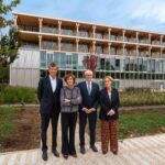 Quatre personnes en tenue professionnelle se tiennent sur un sentier devant un immeuble moderne de plusieurs étages, construit en verre et en bois, entouré d’espaces verts aménagés et d’arbres, sous un ciel partiellement nuageux.