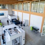 Un vaste espace de travail de style industriel, équipé de grosses machines, avec de hauts plafonds, des bureaux aux parois vitrées et plusieurs personnes qui s'y promènent ou y travaillent. La lumière du soleil pénètre à flots par les hautes fenêtres, et le sol est en béton poli.