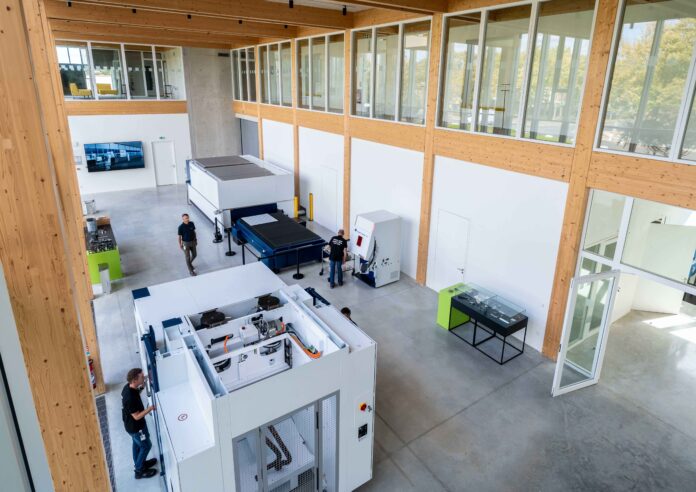 Un vaste espace de travail de style industriel, équipé de grosses machines, avec de hauts plafonds, des bureaux aux parois vitrées et plusieurs personnes qui s'y promènent ou y travaillent. La lumière du soleil pénètre à flots par les hautes fenêtres, et le sol est en béton poli.
